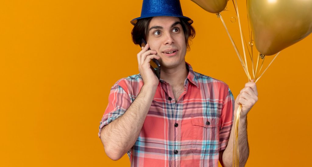 bonito-homem-caucasiano-impressionado-com-chapeu-de-festa-azul-segurando-baloes-de-helio-falando-ao-telefone_141793-99983